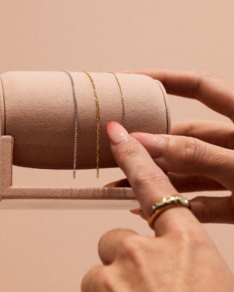 Ein Finger zeigt auf eine Auswahl verschiedener filigraner Ketten, die für ein Permanent Bracelet zur Verfügung stehen. Die Ketten sind in Gold- und Weißgold-Optionen erhältlich und variieren in Design und Struktur.