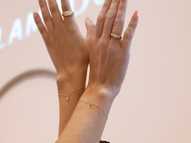 Zwei Hände, die in die Luft gehalten werden, mit permanenten Armbändern an den Handgelenken. Die Armbänder sind filigran und tragen kleine Anhänger, die dezent glänzen.